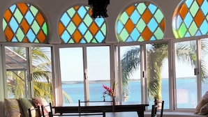 Uma área de refeições com mesa e cadeiras, janelas amplas com padrões coloridos e vista para o mar.