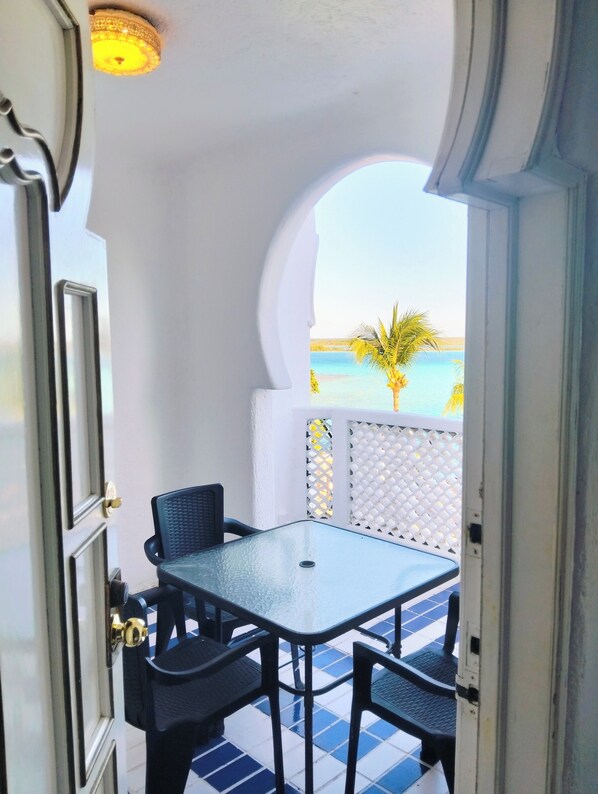 A room with a table and chairs, a view of the sea, and a palm tree.