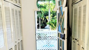 Um corredor com piso de azulejos brancos, uma porta decorativa com padrão geométrico e vista para o verde lá fora.