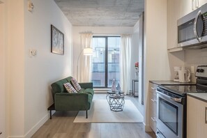 A modern apartment with a green sofa, a dining table with a vase, a kitchen with a microwave and stove, and a large window with curtains. - Central -Parking- 100 walkscore (Montreal)