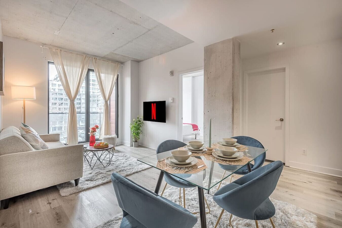 A modern living room with a sofa, dining table, chairs, a TV mounted on the wall, and a large window with curtains.