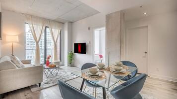 A modern living room with a sofa, dining table, chairs, a TV mounted on the wall, and a large window with curtains.