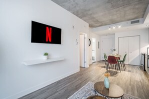 Un espai modern d'estar amb un televisor penjat que mostra una 'N' vermella, una zona de menjador amb taula i cadires, i una petita planta sobre una estant.