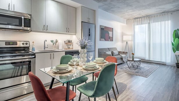 Une cuisine moderne équipée d’appareils électroménagers en acier inoxydable, un coin repas avec une table et des chaises en verre, et un salon avec un canapé et une grande fenêtre.