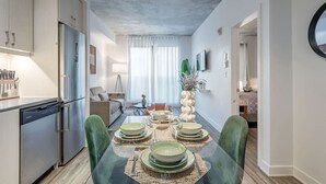Une cuisine moderne équipée d’appareils électroménagers en acier inoxydable, un coin repas avec une table en verre et des chaises vertes, et un salon avec un canapé et un téléviseur.