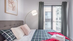 Hotellihuone, jossa on sänky, lattiavalaisin, ikkuna verhoilla ja seinällä oleva kehystetty taulu.