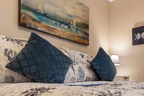 A bed with a patterned bedspread and two dark blue pillows, a painting of a coastal scene on the wall, and a nightstand with a lamp. - Ocean view Retreat 1BR Condo (Murrells Inlet)