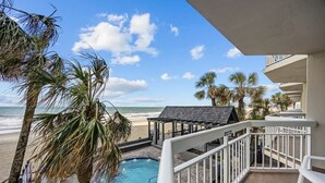 A balcony with a pool, palm trees, and a view of the beach and ocean. - The Tide Calls 1BR Ocean Front (Murrells Inlet)
