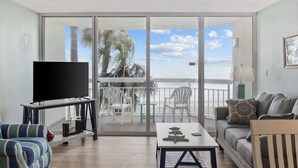 A living room with a TV, a sofa, a coffee table, and a balcony with chairs and a view of the ocean. - The Tide Calls 1BR Ocean Front (Murrells Inlet)