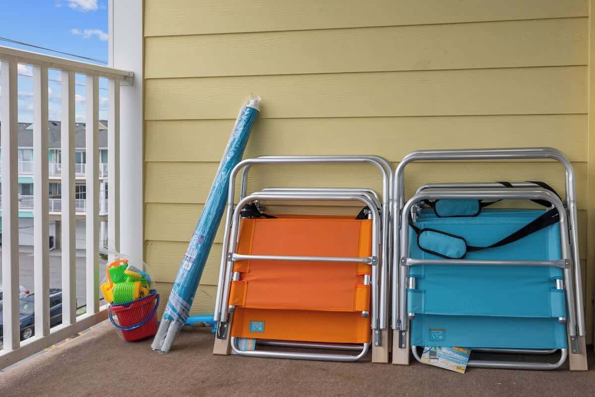 A balcony with two metal folding chairs, one orange and one blue, and a blue mat leaning against the wall.