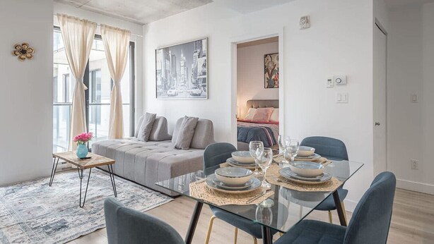 A modern living room with a glass dining table, grey sofa, and a cityscape painting. - Square Vic | 2 Queens + Balcony Retreat (Montreal)
