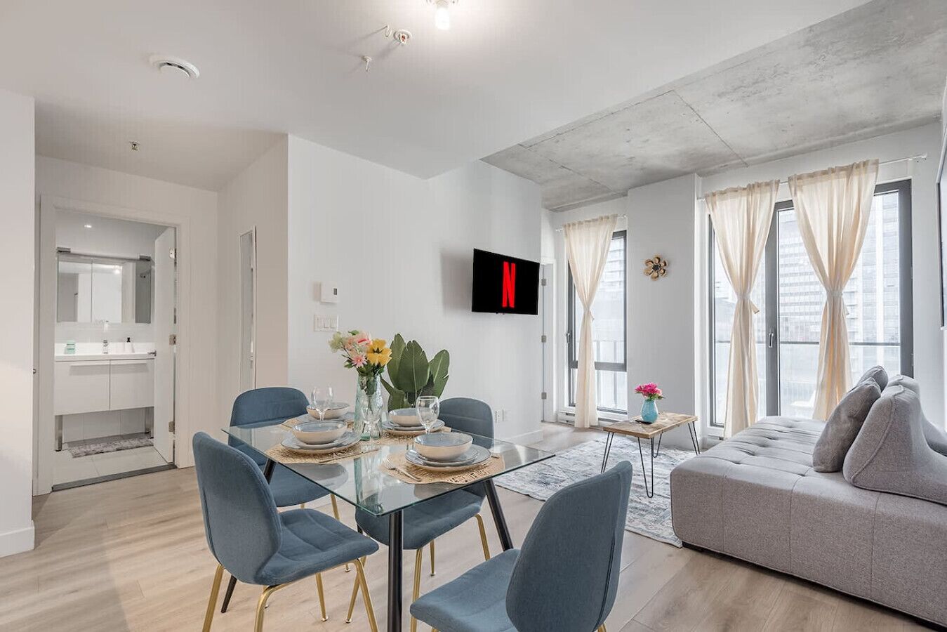 Un salon moderne avec une table à manger en verre, des chaises bleues, un canapé gris et un téléviseur fixé au mur.