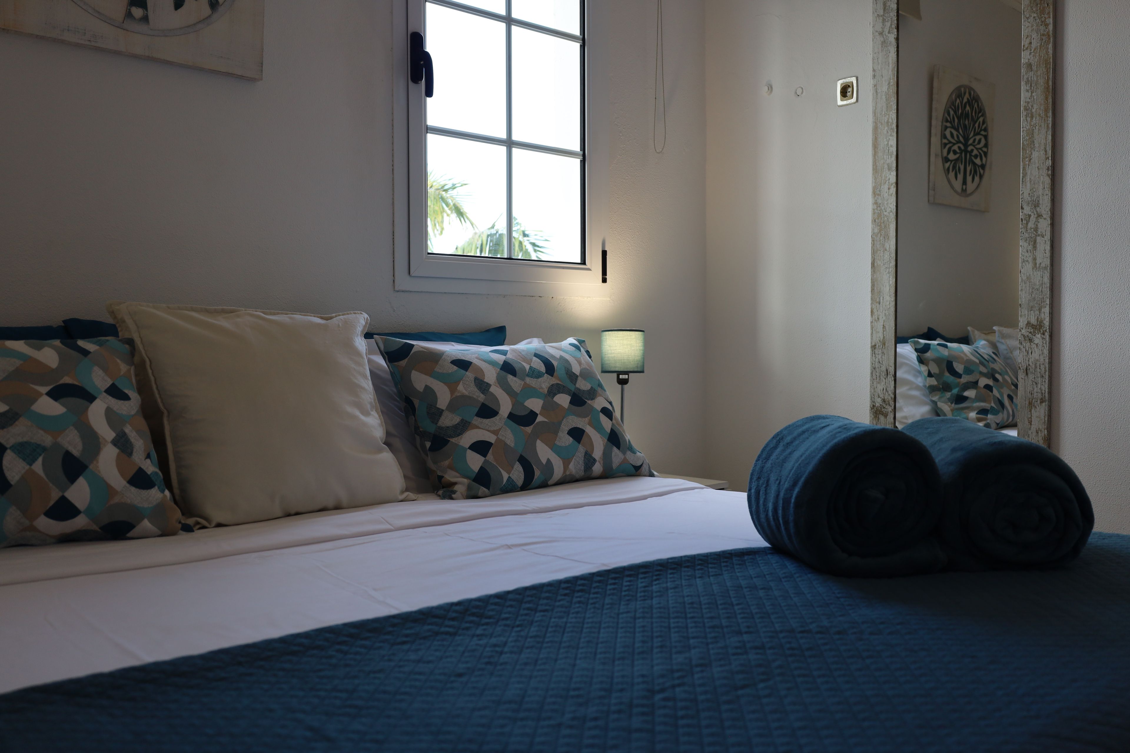 A bed with patterned pillows, a bedside lamp, and a window with a view of palm trees.