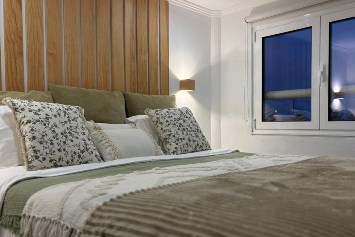 Un dormitorio con una cama, almohadas, una lámpara de noche y una ventana.