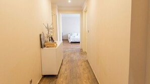 A narrow hallway with a wooden floor leading to a bedroom with a white bed. - Morning in Prague – Cozy Entire Home for 15, 1 min to Tram, 4 Rooms & 2 Kitchens (Praha2)
