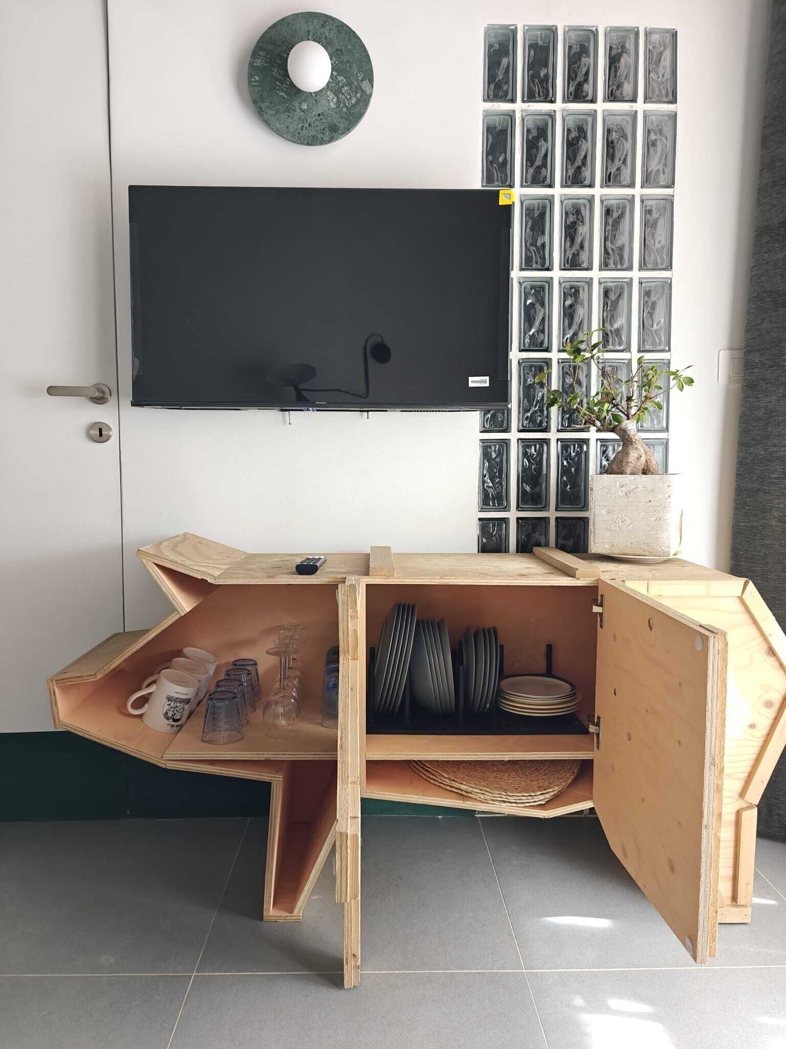 Una stanza con TV a parete, un mobiletto in legno con una pianta e una finestra con quadri.