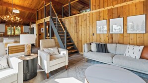 A spacious living area with a staircase, wooden walls, a sofa, and a coffee table. - The Lakeside Lookout - Lakefront 4BR Cabin - Dock, Kayaks, Swim Raft + Game Shed (Cushing)