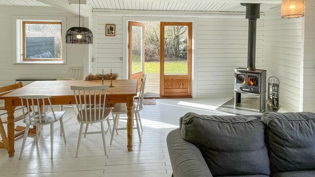 A cozy living room with a fireplace, a dining area with a wooden table and chairs, and a large window with a view of the outdoors. - 3 bedroom lovely home in Sjællands Odde (Sjællands Odde)