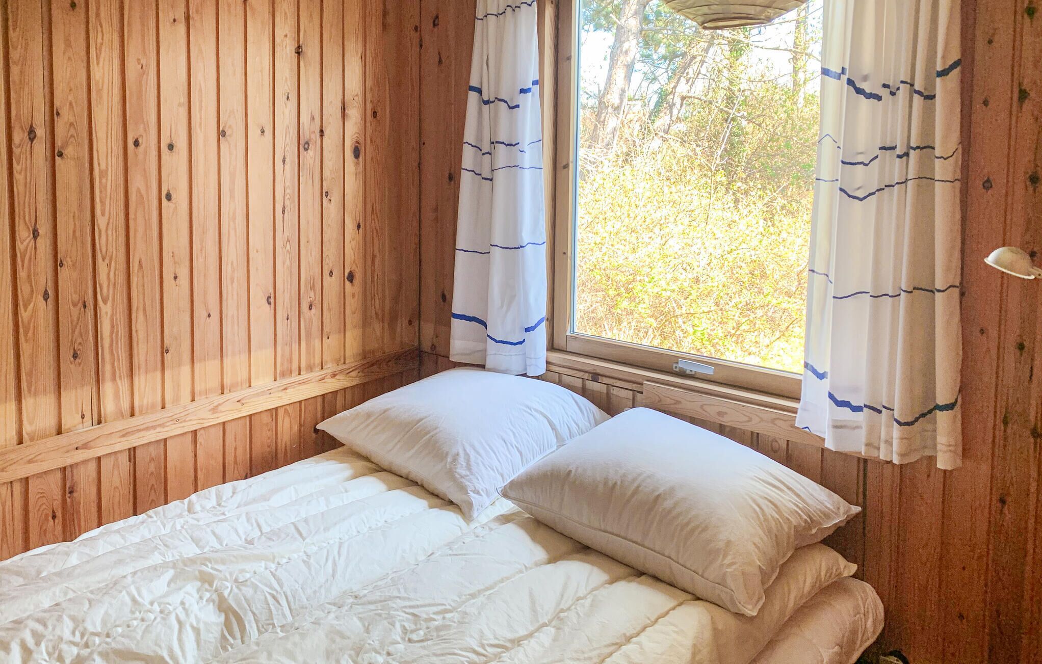 Une chambre dans un chalet en bois, avec un lit, deux oreillers et une fenêtre avec des rideaux.
