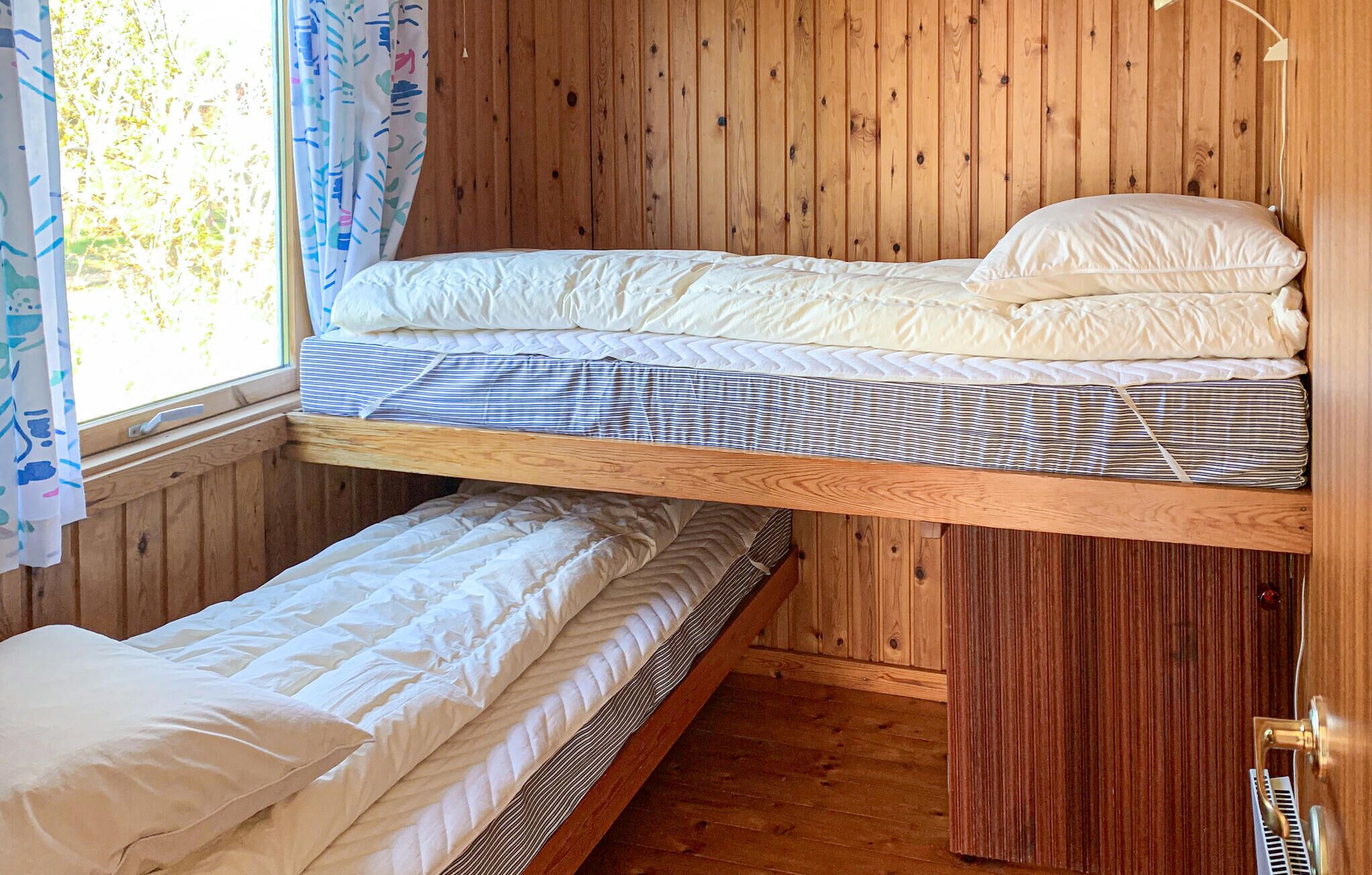 A bunk bed room with wooden walls, a window with curtains, and a bed with a striped mattress.