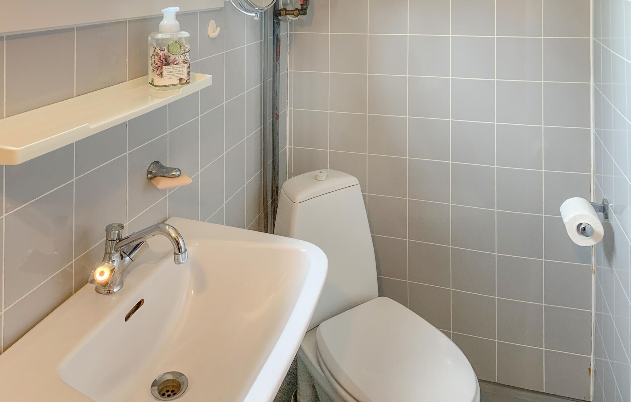 Un baño con lavamanos, inodoro y ducha blancos. Hay un dispensador de jabón en un estante sobre el lavamanos.