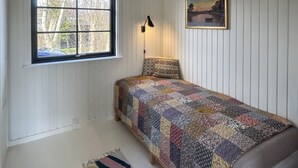 Et soveværelse med vindue, seng med mønstret dyne, lampe og et maleri på væggen.