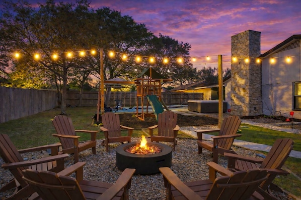 Un patio trasero con un pozo de fuego, sillas Adirondack, un columpio y luces colgantes.