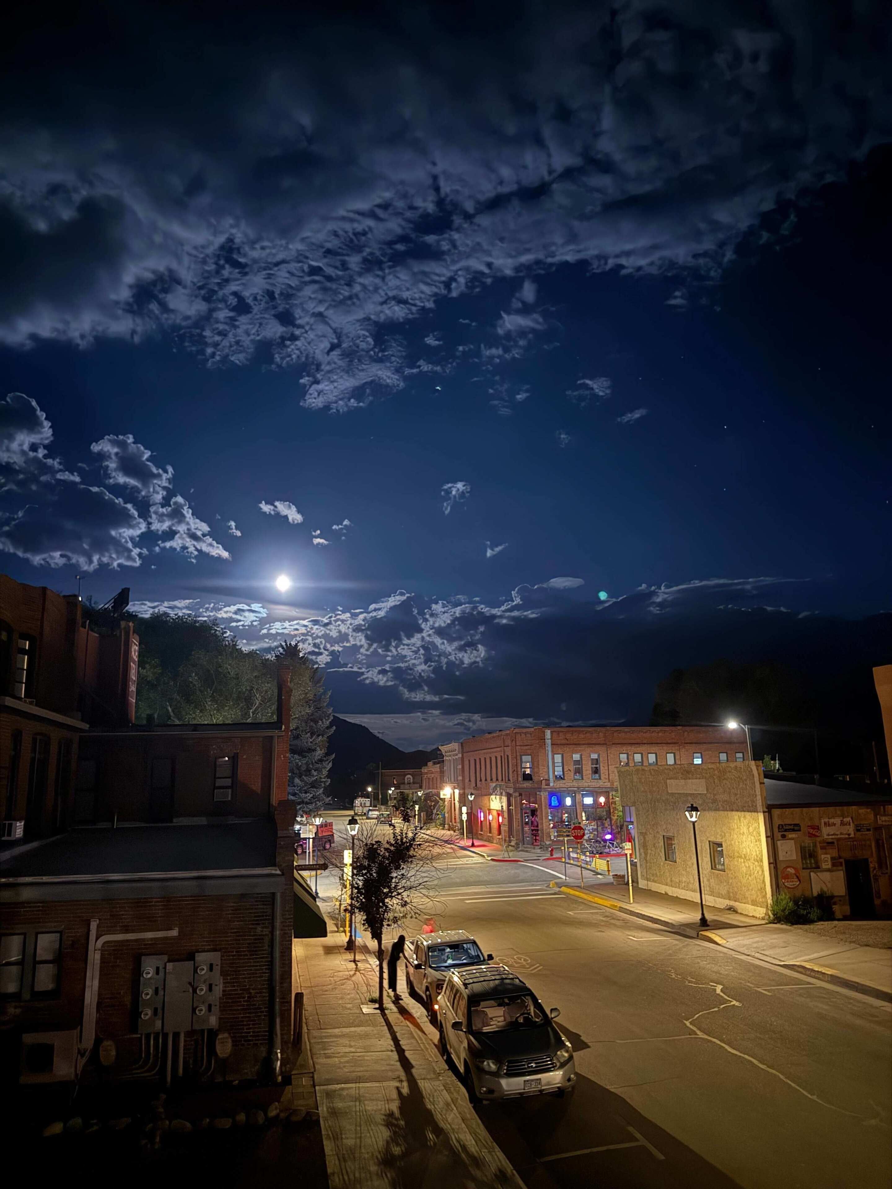 A nighttime street scene with buildings, a car, and a visible moon.