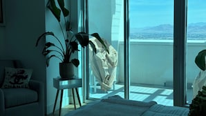 Camera da letto con un letto, una pianta in vaso, una sedia e vista sulla città attraverso una porta scorrevole in vetro.