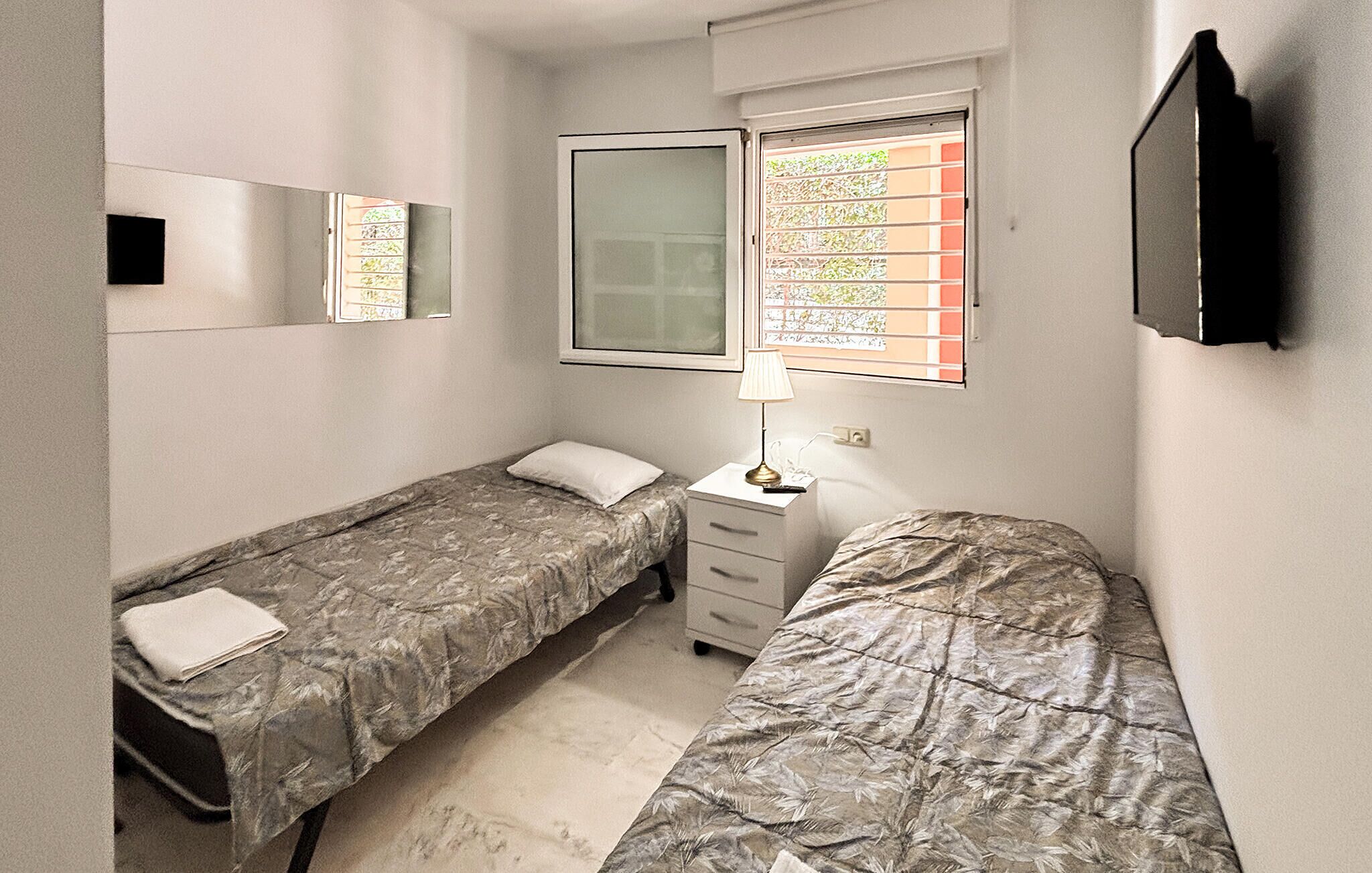 Ein kleines Einzelzimmer mit zwei Einzelbetten, einem Nachttisch mit Lampe, einem Fenster mit Jalousien, einem Spiegel und einem an der Wand befestigten Fernseher.