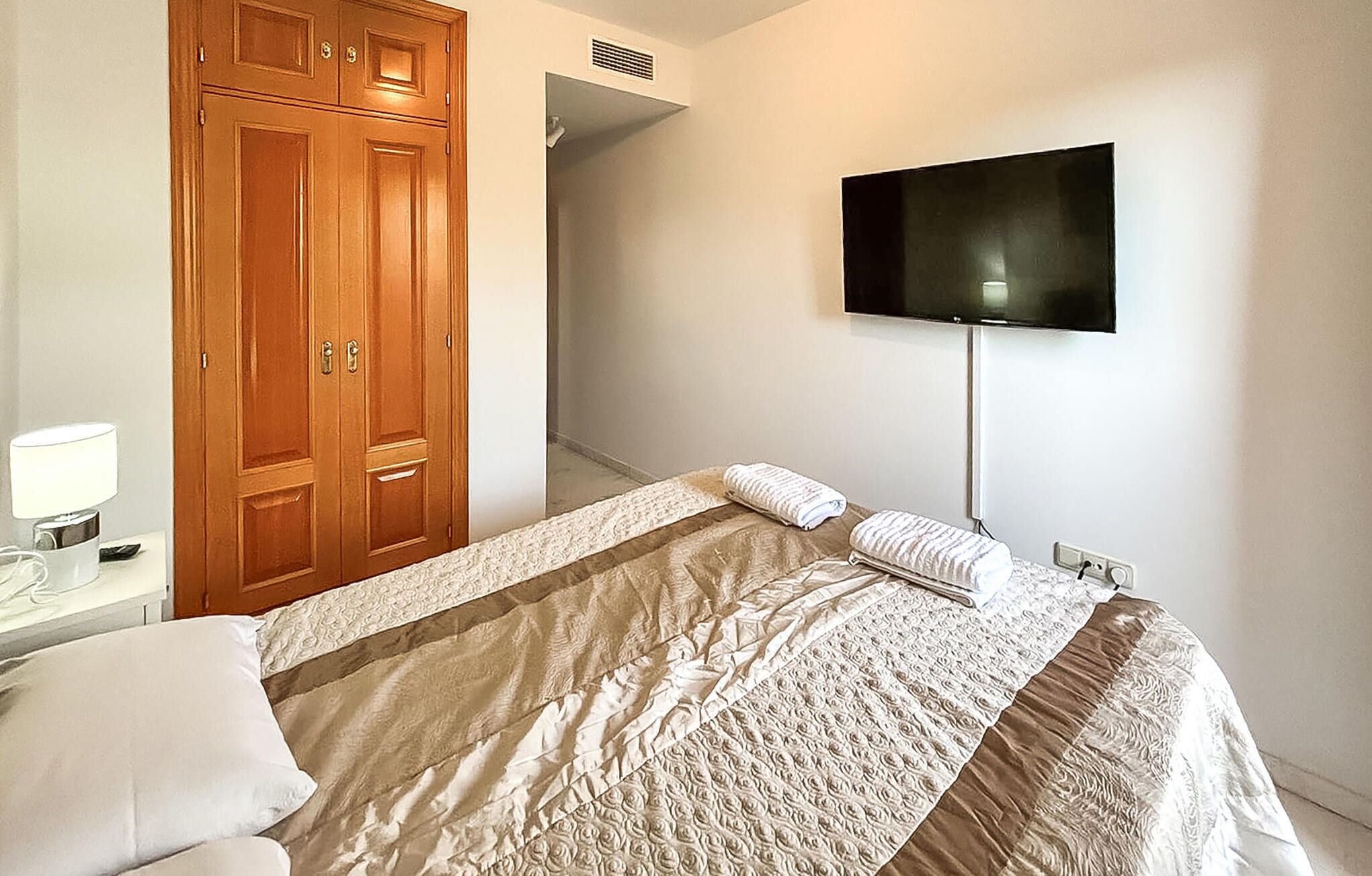 Un dormitorio con una cama, televisión montada en la pared, armario de madera y mesita de noche con lámpara.