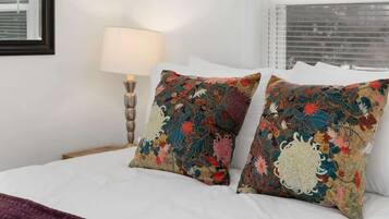 Un lit avec du linge de lit blanc, deux coussins décoratifs, un plaid violet et une lampe de chevet.