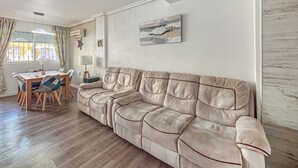 Una sala de estar con un sofá color beige, una mesa de comedor de madera, sillas y una ventana con cortinas.