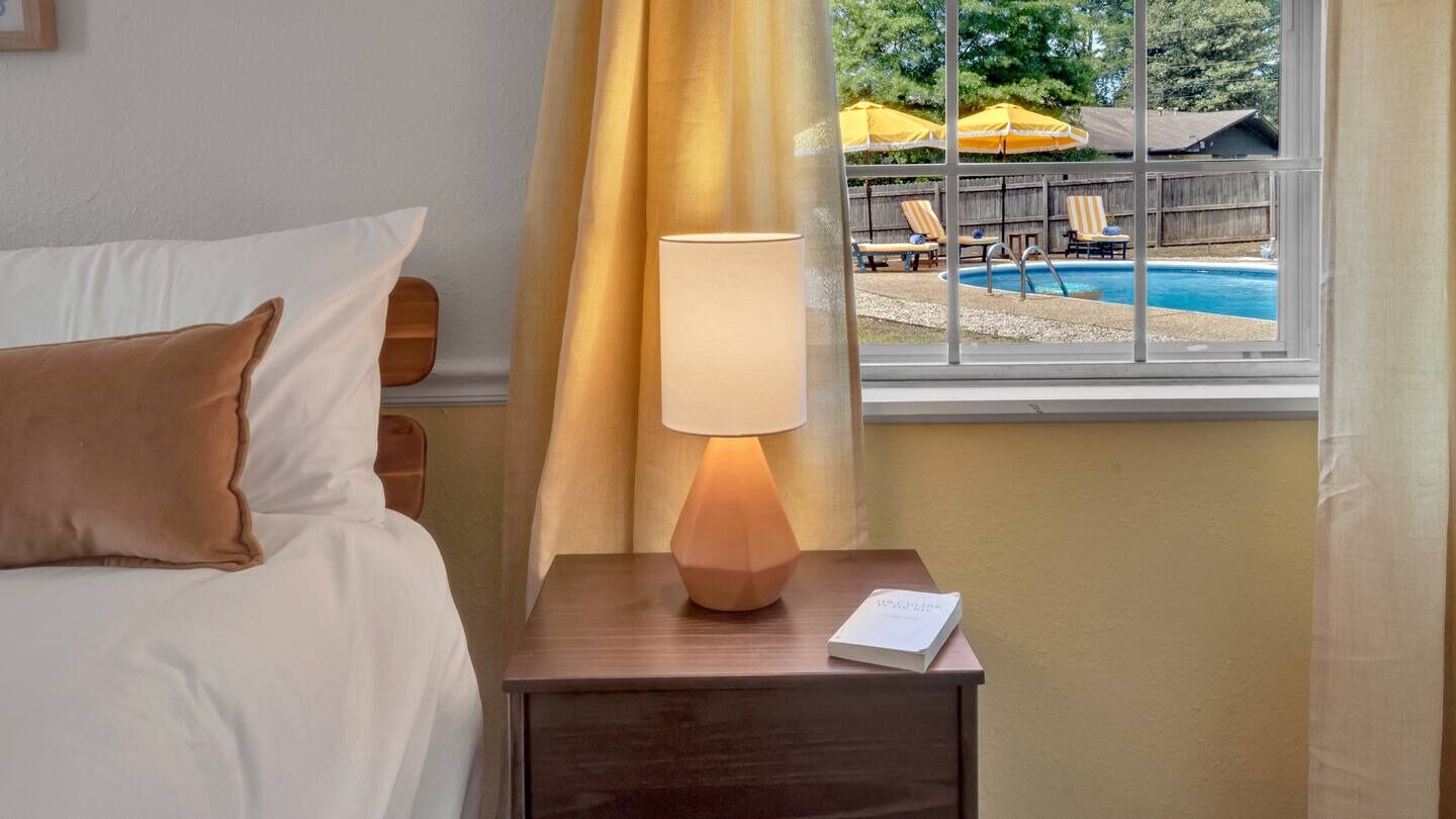 A hotel room with a bed, a bedside table with a lamp, and a view of a pool area with umbrellas and lounge chairs.