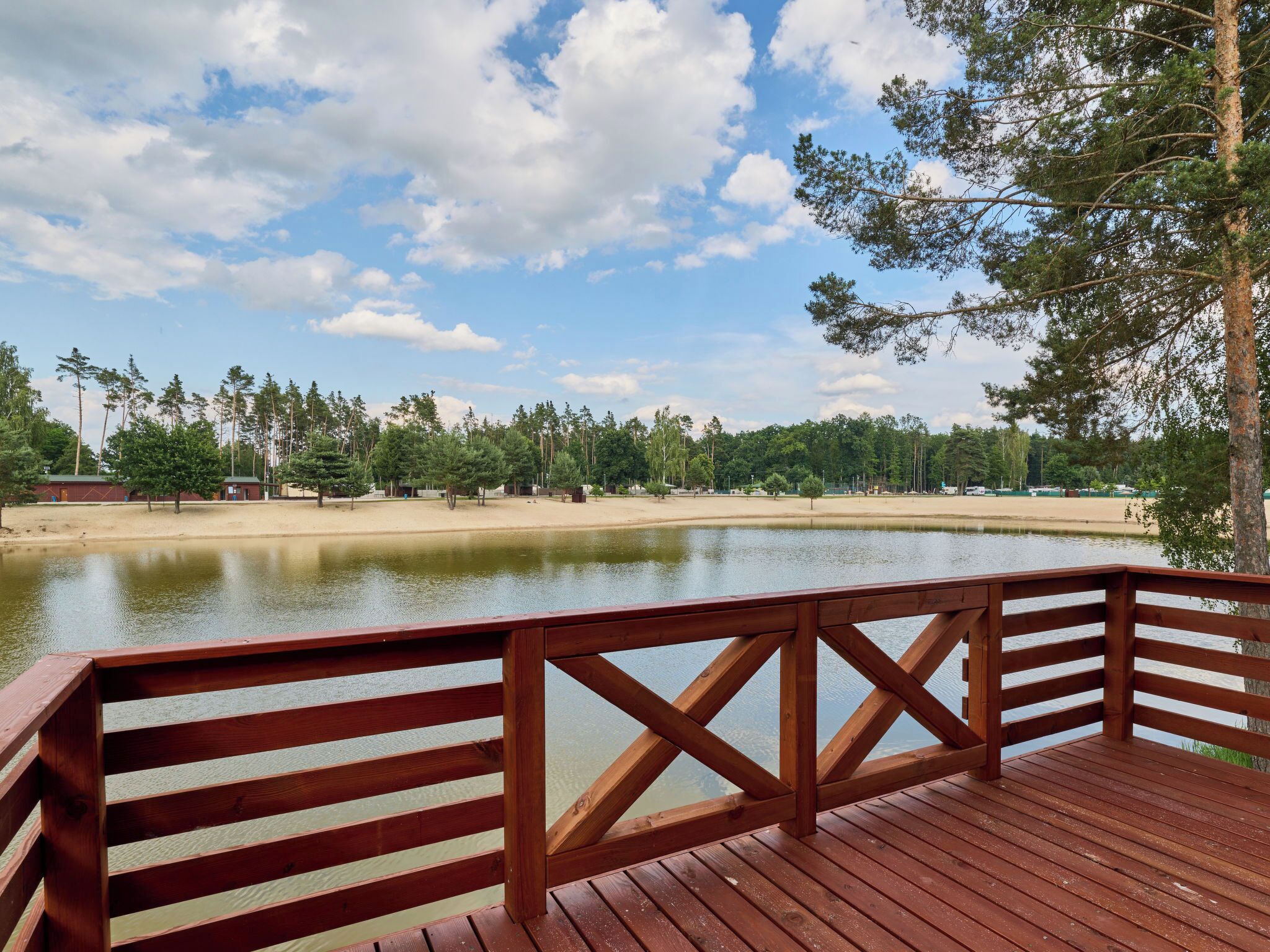 Une terrasse en bois donnant sur un lac, avec une plage de sable et des arbres en arrière-plan.