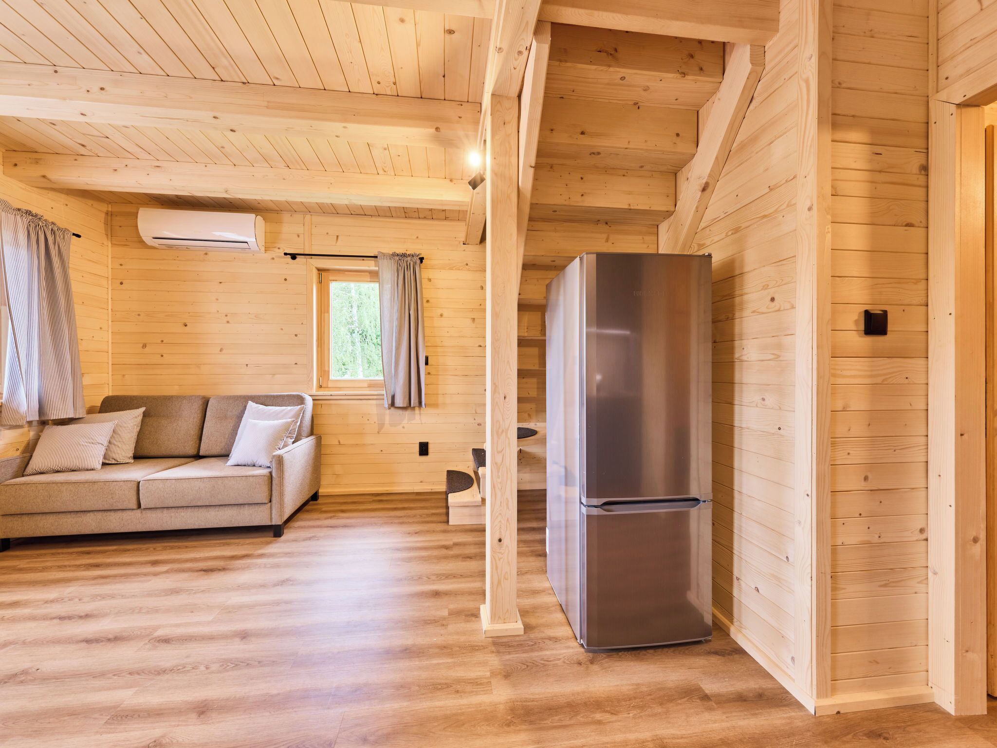 A wooden cabin interior with a sofa, a refrigerator, and a window with curtains.