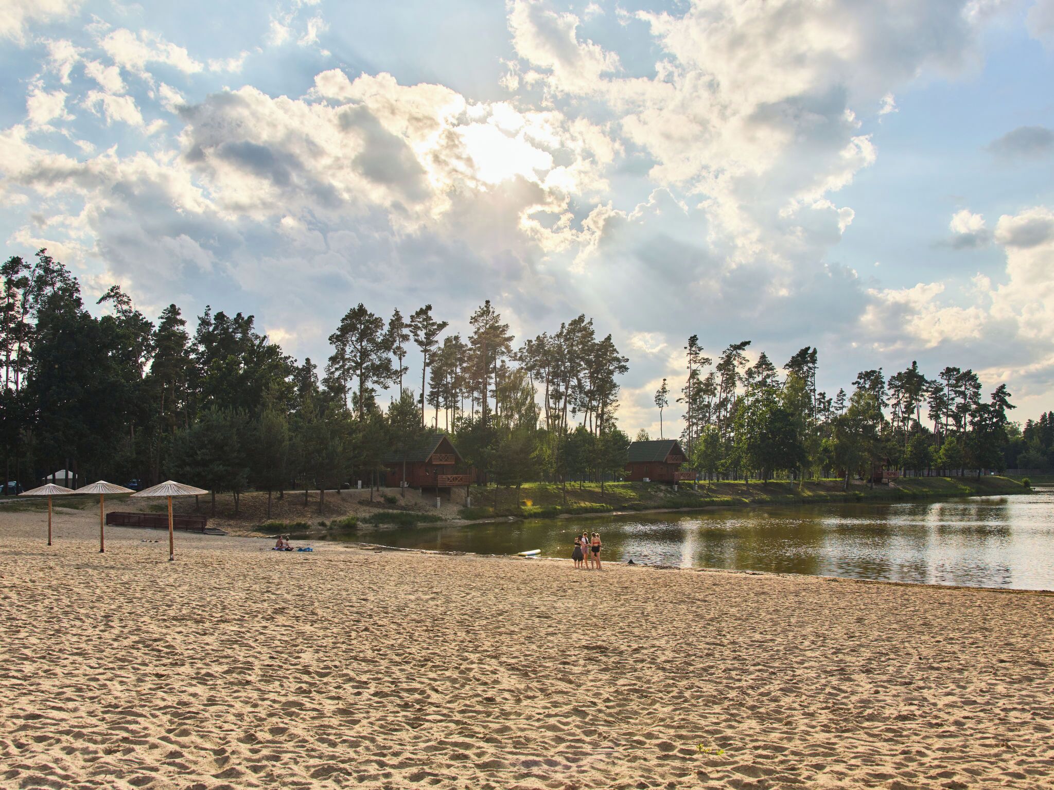 A beach with sand, umbrellas, a lake, and trees in the background.