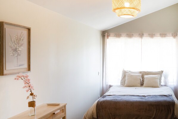Un dormitorio con cama, mesita de noche, un jarrón con flores, un cuadro en la pared y una ventana con cortinas.