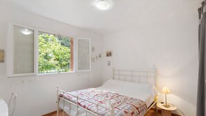 Un dormitorio con cama, lámpara de noche, ventana con vistas a zonas verdes y una pequeña mesa.
