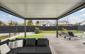 Un espace extérieur abrité comprenant un canapé noir, une petite pelouse et une piscine avec des chaises longues.