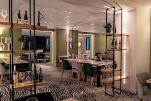 Un comedor moderno con mesas, sillas y botellas de vino. Se observan estantes con plantas y botellas, además de una alfombra estampada.