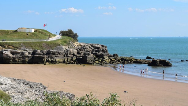 Un paysage côtier avec une plage de sable, des falaises rocheuses et un petit bâtiment situé sur une colline.