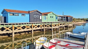 Une rangée de bâtiments en bois aux couleurs vives, avec une jetée en bois qui s’avance sur une étendue d’eau calme, et des bateaux amarrés à la jetée.