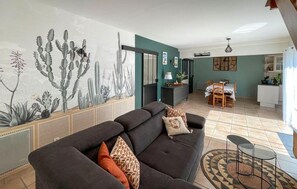 Ein Wohnzimmer mit einem dunklen Sofa, gemusterten Kissen, einer Wandmalerei mit Kakteen, einem Essbereich mit Tisch und Stühlen sowie einer Küchenzeile inklusive Kühlschrank.