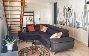 Ein Wohnzimmer mit einem Ecksofa, einem kleinen runden Tisch, einem Fernseher, einer Wandleiter und einer Kakteen-Wandkunst.