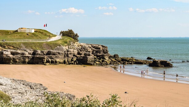 Paesaggio costiero con spiaggia sabbiosa, scogliere rocciose e un pendio erboso con una bandiera.