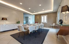Ein Essbereich mit einem langen Tisch, gedeckt für sechs Personen, weißen Stühlen, einer Glastrennwand und einer modernen Pendelleuchte.