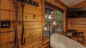 Interior de una cabaña de madera con una silla blanca, una ventana con vistas y un televisor montado.