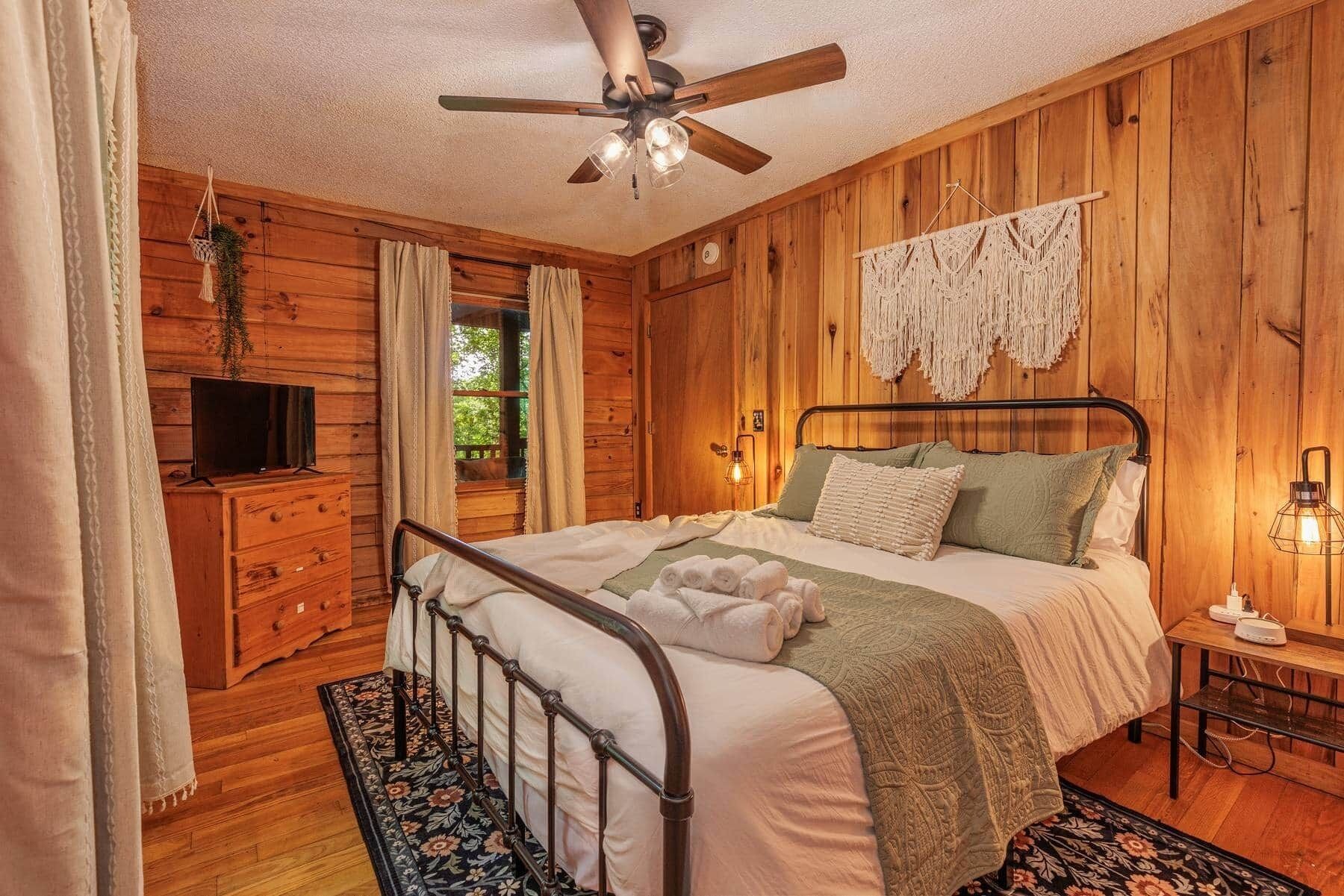 A wooden-paneled bedroom with a bed, a ceiling fan, a TV, and a bedside table with a lamp.