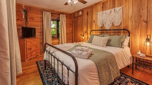Un dormitorio con paneles de madera, una cama, un ventilador de techo, un televisor y una mesita de noche con una lámpara.